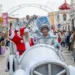 Babbo Natale e una persona con un costume d'argento lucido viaggiano su un'auto bianca durante la parata di Natale a Loulé in Algarve, circondati dalla folla e da bambini che guardano e scattano foto. Decorazioni ed edifici fanno da sfondo.
