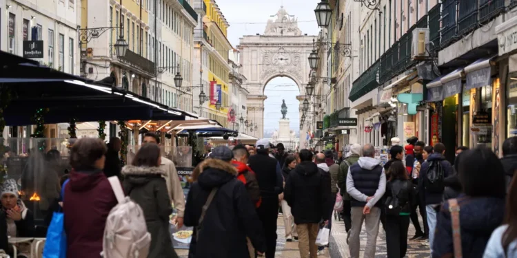 Una trafficata strada pedonale di Lisbona in Portogallo, fiancheggiata da negozi e caffè e affollata di persone creata dall'economia locale di quest'anno. Sullo sfondo spiccano un grande arco decorativo e una statua, che riflettono l'atmosfera vivace e urbana.