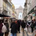 Una trafficata strada pedonale di Lisbona in Portogallo, fiancheggiata da negozi e caffè e affollata di persone creata dall'economia locale di quest'anno. Sullo sfondo spiccano un grande arco decorativo e una statua, che riflettono l'atmosfera vivace e urbana.
