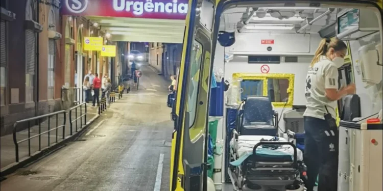Un'ambulanza con le porte aperte è parcheggiata di notte davanti all'ingresso di un ospedale, dove brilla il cartello Pronto soccorso chiuso. Un paramedico organizza l'attrezzatura all'interno, mentre sullo sfondo sono visibili alcune persone in strada.