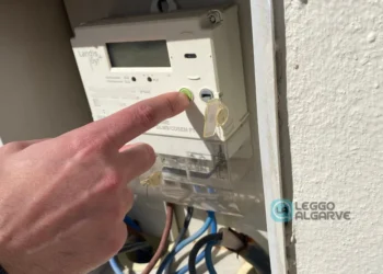 La mano di una persona preme un pulsante verde su un contatore elettrico installato su una parete, con i fili visibili sotto. Il contatore aperto accenna alla verifica dei costi dell'energia elettrica mentre la luce del sole proietta ombre sulla superficie.