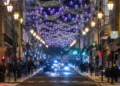 Una strada di città di notte decorata con luci viola a forma di stella e ghirlande in alto, fiancheggiata da lampioni luminosi e affollata di persone - portoghesi quanto spendono Natale - che si godono lo shopping natalizio mentre le auto percorrono la strada.
