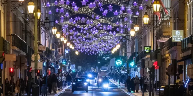Una strada di città di notte decorata con luci viola a forma di stella e ghirlande in alto, fiancheggiata da lampioni luminosi e affollata di persone - portoghesi quanto spendono Natale - che si godono lo shopping natalizio mentre le auto percorrono la strada.