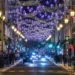 Una strada di città di notte decorata con luci viola a forma di stella e ghirlande in alto, fiancheggiata da lampioni luminosi e affollata di persone - portoghesi quanto spendono Natale - che si godono lo shopping natalizio mentre le auto percorrono la strada.
