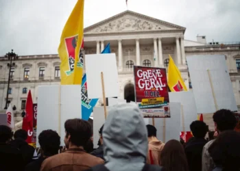 Un gruppo di persone di spalle protestano davanti al parlamento in Portogallo, per lo sciopero generale. Hanno bandiere gialle e rosse e cartelli con scritto Greve Geral.
