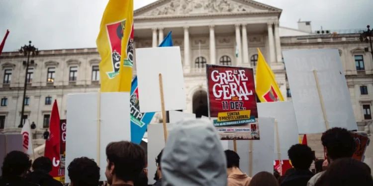 Un gruppo di persone di spalle protestano davanti al parlamento in Portogallo, per lo sciopero generale. Hanno bandiere gialle e rosse e cartelli con scritto Greve Geral.
