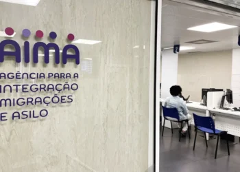 Una persona siede in un'area d'attesa dell'ufficio dell'Agência para a Integração Migrações e Asilo (AIMA), in attesa del proprio turno per l'aggiornamento dati utenti AIMA, con banchi di servizio numerati e una parete di vetro con il nome e il logo dell'agenzia.
