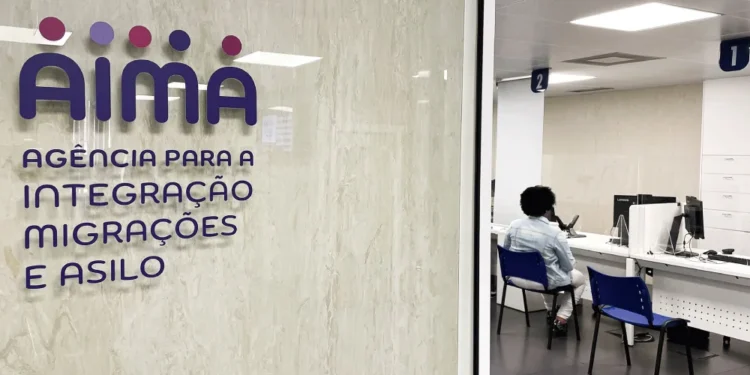 Una persona siede in un'area d'attesa dell'ufficio dell'Agência para a Integração Migrações e Asilo (AIMA), in attesa del proprio turno per l'aggiornamento dati utenti AIMA, con banchi di servizio numerati e una parete di vetro con il nome e il logo dell'agenzia.