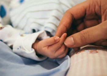 Primo piano di una mano adulta che stringe delicatamente la manina di un neonato. Il bambino, con un morbido vestitino a righe, incarna calore e cura: una scena che riecheggia lo spirito del recente ongedo parentale dal lavoro in Portogallo: le novità.