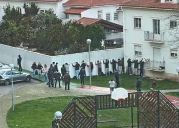 Un gruppo di persone si trova di fronte a un muro bianco con le mani alzate, come in un confronto con la polizia, mentre la GNR blocca la Baby Gang in un'area residenziale con erba, un canestro da basket ed edifici bianchi nelle vicinanze. Altre persone osservano la scena.