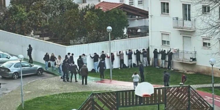 Un gruppo di persone si trova di fronte a un muro bianco con le mani alzate, come in un confronto con la polizia, mentre la GNR blocca la Baby Gang in un'area residenziale con erba, un canestro da basket ed edifici bianchi nelle vicinanze. Altre persone osservano la scena.