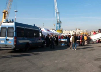 L'arrivo di migranti al porto italiano con un furgone della Polizia per accogliere, si vedono delle gru sul fondo di un cielo azzurro, foto a corredo dell'articolo che parla del Portogallo che paga per non accogliere.