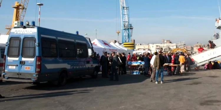 L'arrivo di migranti al porto italiano con un furgone della Polizia per accogliere, si vedono delle gru sul fondo di un cielo azzurro, foto a corredo dell'articolo che parla del Portogallo che paga per non accogliere.