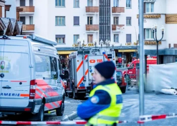 La polizia e i veicoli di emergenza sono parcheggiati in una strada di fronte a un edificio a Crans-Montana. Un agente offuscato si trova in primo piano, con parti dell'area transennate in seguito alle indagini sulle vittime portoghesi.