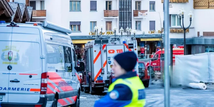 La polizia e i veicoli di emergenza sono parcheggiati in una strada di fronte a un edificio a Crans-Montana. Un agente offuscato si trova in primo piano, con parti dell'area transennate in seguito alle indagini sulle vittime portoghesi.