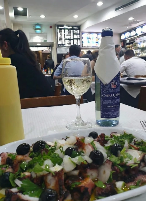 Un piatto tipico di polpo con olive e prezzemolo accanto a una bottiglia di vino bianco su un tavolo da ristorante, rappresentando l'eccellenza culinaria lusitana nel difficile contesto della crisi ristoranti portoghesi.