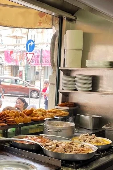 Una cucina della tradizione di un ristorante portoghese con alcune padelle con bifanas in vetrina e dei salgados. Sullo sfondo si vede l'esterno del locale: una strada con un'auto e alcune persone.
