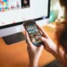Una persona utilizza uno smartphone per sfogliare una galleria fotografica mentre è seduta a una scrivania con monitor, tastiera e un piccolo cactus in vaso, magari riflettendo su notizie come "Portogallo vieta social minori 16 anni.