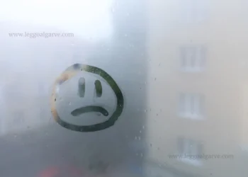 Un volto triste è disegnato su una finestra nebbiosa, con edifici sfocati visibili all'esterno, catturando lo stato d'animo di depressione di Portogallo. Il logo "LEGGO ALGARVE" appare in basso a destra.