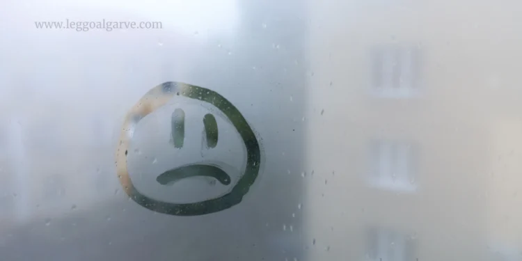 Un volto triste è disegnato su una finestra nebbiosa, con edifici sfocati visibili all'esterno, catturando lo stato d'animo di depressione di Portogallo. Il logo "LEGGO ALGARVE" appare in basso a destra.