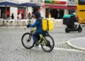 Un corriere per le consegne di cibo che indossa una giacca blu e uno zaino giallo attraversa in bicicletta una strada acciottolata, mentre sullo sfondo passa un altro corriere in scooter con uno zaino verde - nonostante le occasionali lamentele del servizio di consegna.