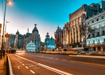 Un'ampia strada cittadina al tramonto con semafori sfocati, edifici storici e una cupola sormontata da una statua, probabilmente in una città europea nota per le discussioni sul salario minimo in Spagna. Il cielo è limpido e azzurro.