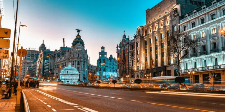 Un'ampia strada cittadina al tramonto con semafori sfocati, edifici storici e una cupola sormontata da una statua, probabilmente in una città europea nota per le discussioni sul salario minimo in Spagna. Il cielo è limpido e azzurro.