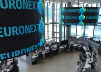 Una moderna sala di negoziazione presso la sede di Euronext, dove diverse persone lavorano alle postazioni informatiche. Grandi schermi digitali visualizzano la parola "EURONEXT" e grafici colorati in una stanza dalle pareti di vetro e molto illuminata.