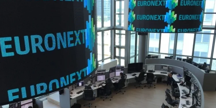 Una moderna sala di negoziazione presso la sede di Euronext, dove diverse persone lavorano alle postazioni informatiche. Grandi schermi digitali visualizzano la parola "EURONEXT" e grafici colorati in una stanza dalle pareti di vetro e molto illuminata.