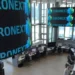 Una moderna sala di negoziazione presso la sede di Euronext, dove diverse persone lavorano alle postazioni informatiche. Grandi schermi digitali visualizzano la parola "EURONEXT" e grafici colorati in una stanza dalle pareti di vetro e molto illuminata.