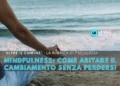Una persona siede a gambe incrociate su una spiaggia in una posizione meditativa con una mano appoggiata sul ginocchio. Il testo in italiano recita: "Mindfulness: come abitare il cambiamento senza perdersi" con il logo di Leggo Algarve.