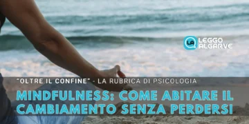 Una persona siede a gambe incrociate su una spiaggia in una posizione meditativa con una mano appoggiata sul ginocchio. Il testo in italiano recita: "Mindfulness: come abitare il cambiamento senza perdersi" con il logo di Leggo Algarve.