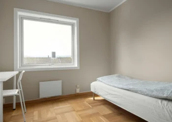 Una camera minimal con pareti beige chiaro, un letto singolo con piumone a pois, una scrivania e una sedia bianche, parquet in legno e un'ampia finestra che lascia entrare la luce naturale: l'ideale per chi vuole affittare stanze Portogallo.