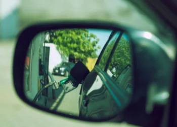 Lo specchietto laterale di un'auto riflette l'immagine di un erogatore verde inserito nel serbatoio di una stazione di servizio, evidenziando in modo sottile la crisi energetica del Portogallo, con alberi e un altro veicolo visibili sullo sfondo.