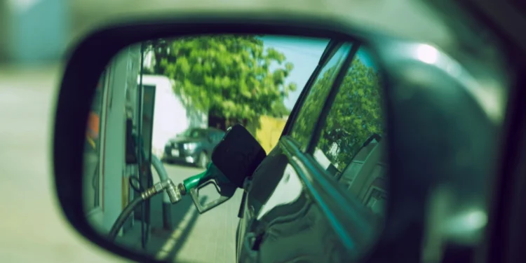 Lo specchietto laterale di un'auto riflette l'immagine di un erogatore verde inserito nel serbatoio di una stazione di servizio, evidenziando in modo sottile la crisi energetica del Portogallo, con alberi e un altro veicolo visibili sullo sfondo.