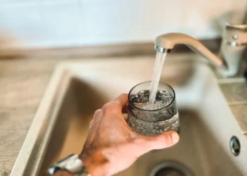 Una mano tiene un bicchiere sotto il rubinetto della cucina in funzione, riempiendolo d'acqua, evidenziando la qualità dell'acqua rubinetto Portogallo. Il lavello e il piano di lavoro sono visibili sullo sfondo.