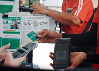 Una persona tiene in mano una carta di pagamento verde mentre un'altra scansiona un codice QR su un dispositivo portatile che offre servizi MB Way Italia; vicino, qualcuno usa uno smartphone. Persone in divisa rossa sono accanto a un tabellone con informazioni e dispositivi.