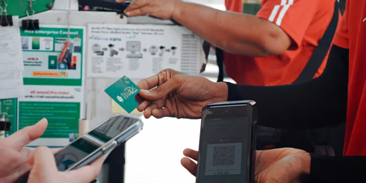 Una persona tiene in mano una carta di pagamento verde mentre un'altra scansiona un codice QR su un dispositivo portatile che offre servizi MB Way Italia; vicino, qualcuno usa uno smartphone. Persone in divisa rossa sono accanto a un tabellone con informazioni e dispositivi.