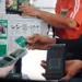 Una persona tiene in mano una carta di pagamento verde mentre un'altra scansiona un codice QR su un dispositivo portatile che offre servizi MB Way Italia; vicino, qualcuno usa uno smartphone. Persone in divisa rossa sono accanto a un tabellone con informazioni e dispositivi.